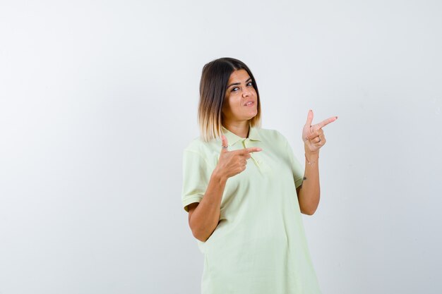 Chica joven apuntando a la derecha con los dedos índices en camiseta y mirando confiado, vista frontal.