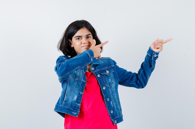 Chica joven apuntando a la derecha con los dedos índices en camiseta roja y chaqueta de jean y mirando feliz. vista frontal.