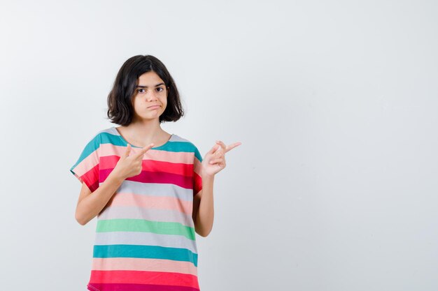 Chica joven apuntando hacia la derecha con los dedos índices, labios curvados en una camiseta a rayas de colores y mirando seriamente, vista frontal.