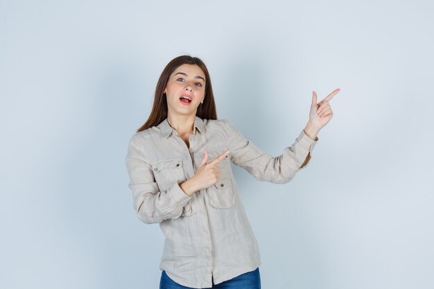 Chica joven apuntando hacia la derecha con los dedos índices, manteniendo la boca abierta en camisa beige, jeans y mirando alegre, vista frontal.