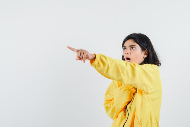 Chica joven apuntando hacia la esquina izquierda con el dedo índice en chaqueta de bombardero amarilla y mirando sorprendido.