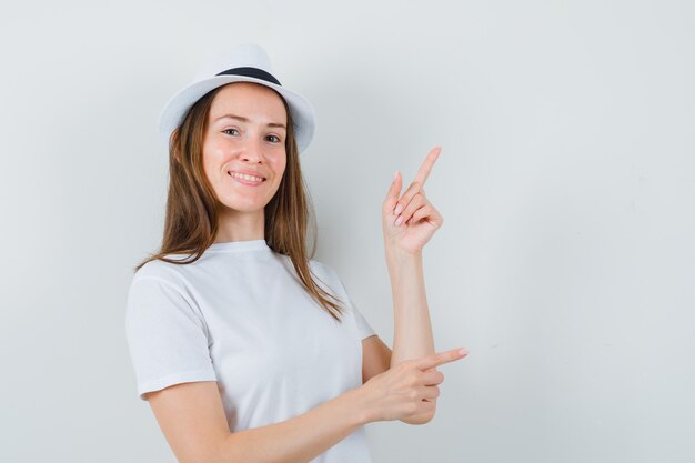 Chica joven apuntando a la esquina superior derecha con camiseta blanca, sombrero y mirando alegre. vista frontal.