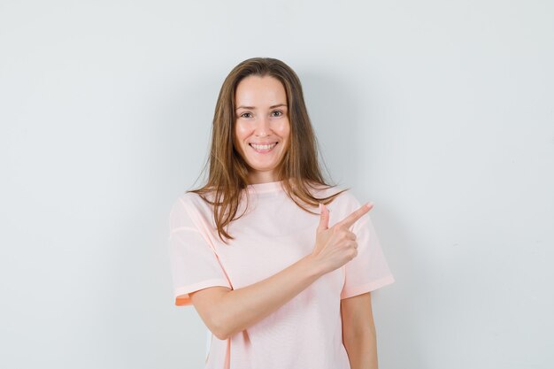 Chica joven apuntando a la esquina superior derecha en camiseta rosa y mirando jovial, vista frontal.