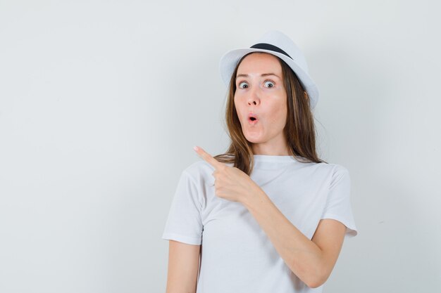 Chica joven apuntando a la esquina superior izquierda con camiseta blanca, sombrero y mirando curioso, vista frontal.