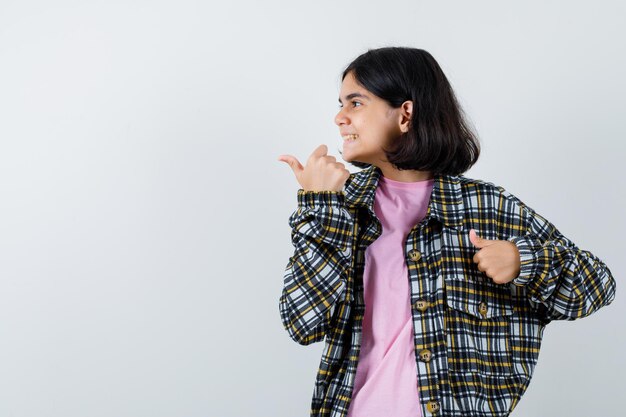 Chica joven apuntando a la izquierda con camisa a cuadros y camiseta rosa y mirando feliz. vista frontal.