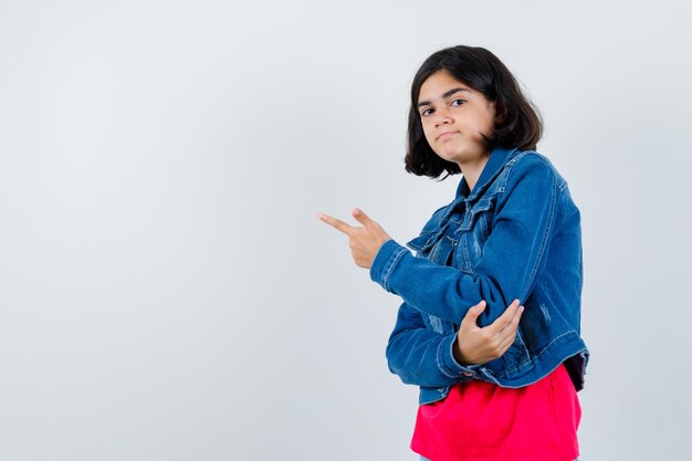 Chica joven apuntando a la izquierda con el dedo índice en camiseta roja y chaqueta de jean y mirando seria. vista frontal.