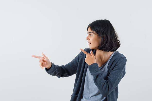 Chica joven apuntando hacia la izquierda con los dedos índices en una camiseta gris claro y una sudadera con capucha frontal con cremallera gris oscuro y mirando feliz