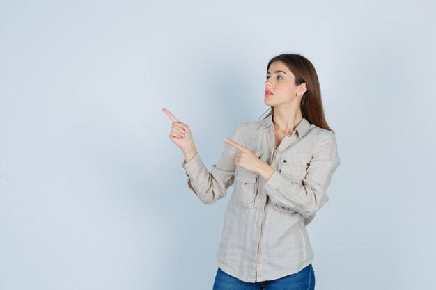 Chica joven apuntando hacia la izquierda con los dedos índices, mirando al lado izquierdo con camisa beige, jeans y mirando enfocado. vista frontal.