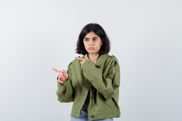 Chica joven apuntando a la izquierda con los dedos índices en suéter gris, chaqueta caqui, pantalón de mezclilla y mirando serio, vista frontal.