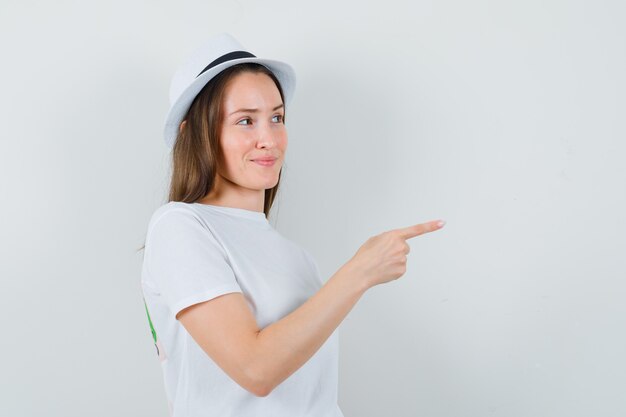 Chica joven apuntando a un lado en camiseta blanca, sombrero y aspecto lindo. vista frontal.