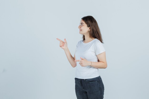 Chica joven apuntando hacia el lado en camiseta, jeans y mirando sensible, vista frontal.