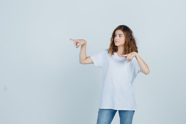 Chica joven apuntando hacia el lado izquierdo en camiseta, jeans y mirando confiado, vista frontal.