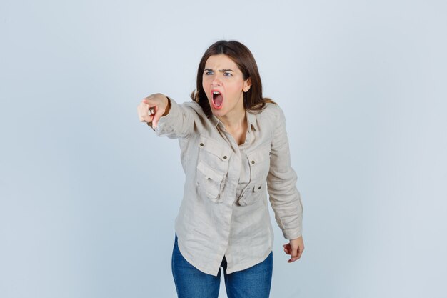 Chica joven apuntando hacia otro lado con el dedo índice de manera enojada en camisa beige, jeans y mirando agitado. vista frontal.