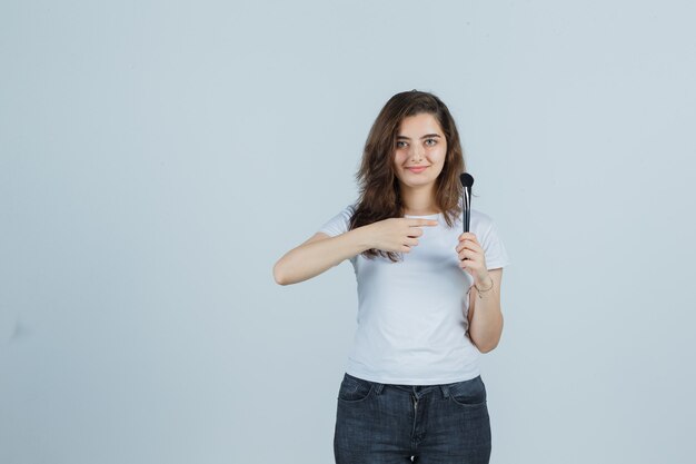 Chica joven apuntando con pincel de maquillaje en camiseta, jeans y mirando confiado, vista frontal.