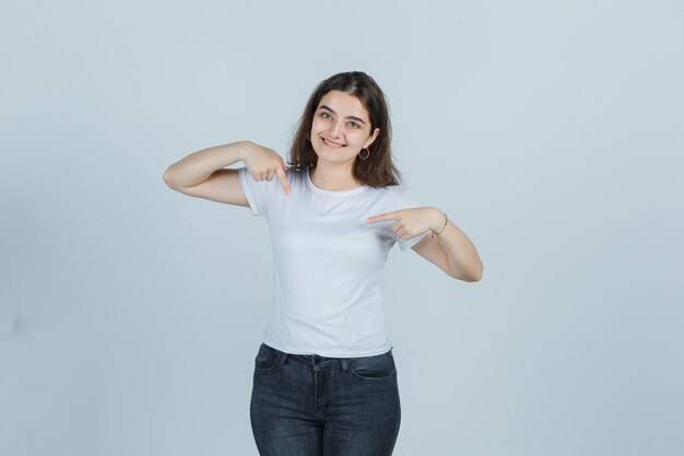 Chica joven apuntando a sí misma en camiseta, jeans y luciendo orgullosa. vista frontal.