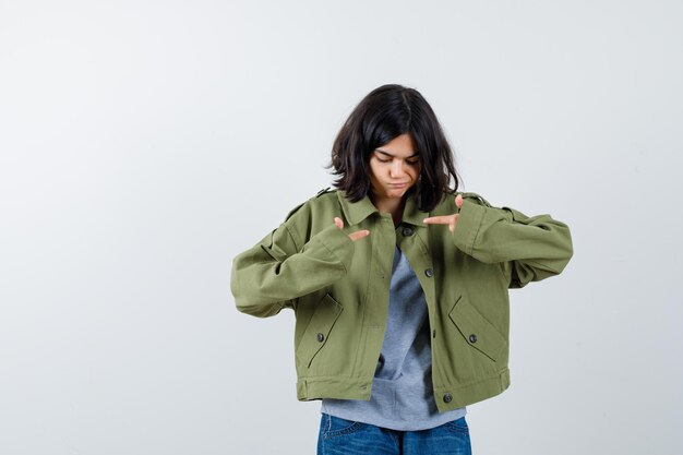 Chica joven apuntando a sí misma en suéter gris, chaqueta caqui, pantalón de mezclilla y mirando enfocado, vista frontal.