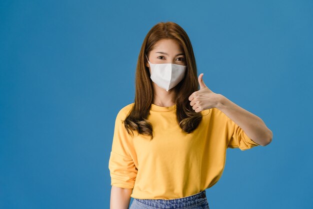 Chica joven de Asia con mascarilla médica mostrando el pulgar hacia arriba vestida con un paño casual y mira a cámara aislada sobre fondo azul. Autoaislamiento, distanciamiento social, cuarentena por coronavirus.