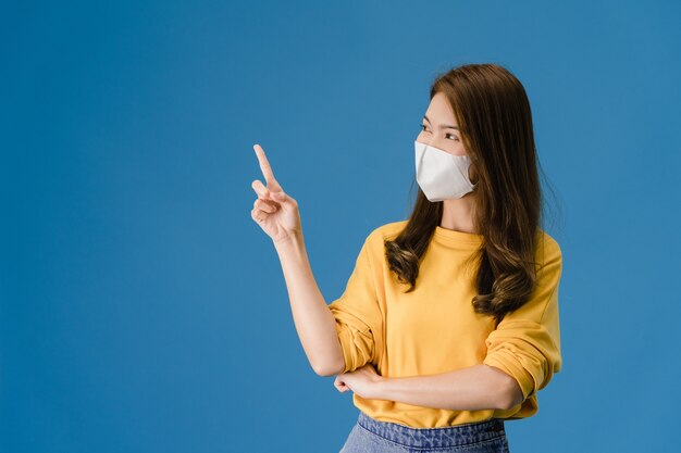 La chica joven de Asia con mascarilla médica muestra algo en el espacio en blanco con vestida con un paño informal y mirando a cámara aislada sobre fondo azul. Distanciamiento social, cuarentena por coronavirus.