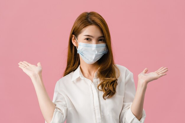 Chica joven de Asia con mascarilla médica que muestra el signo de la paz, anima con vestida con ropa informal y mirando a cámara aislada sobre fondo rosa