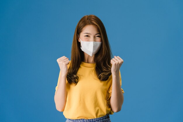 Chica joven de Asia con mascarilla médica que muestra el signo de la paz, animar con vestida con ropa informal y mirando a cámara aislada sobre fondo azul. Distanciamiento social, cuarentena por coronavirus.