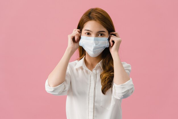 Chica joven de Asia con mascarilla médica vestida con ropa casual y mirando a cámara aislada sobre fondo rosa.