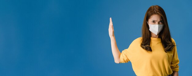 Chica joven de Asia usa mascarilla haciendo dejar de cantar con la palma de la mano con expresión negativa y mirando a la cámara. Distanciamiento social, cuarentena por coronavirus. Bandera panorámica de fondo azul.