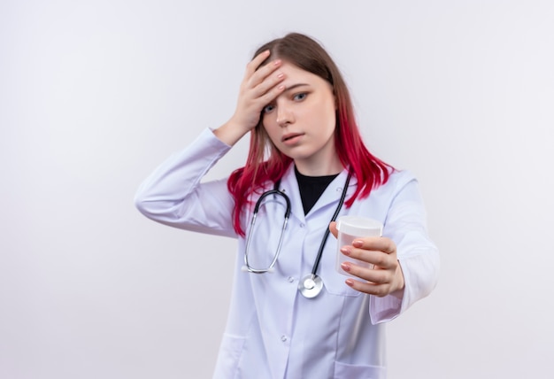 Chica joven asustada con estetoscopio bata médica mirando lata vacía en su mano y poniendo la mano en la frente sobre fondo blanco aislado