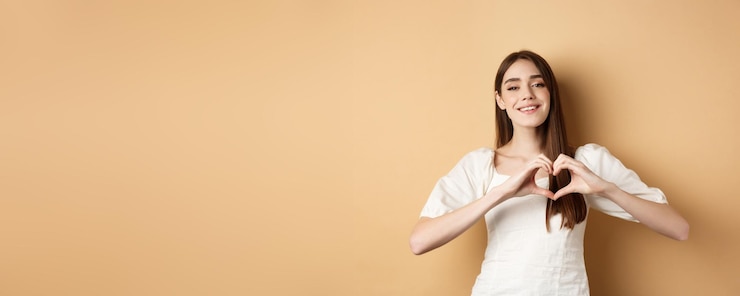 Foto gratuito chica joven atractiva del día de san valentín en vestido blanco sonriendo y mostrando gesto de corazón de pie en ser