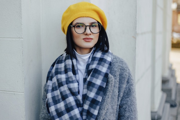 Chica joven atractiva con gafas en abrigo y boina amarilla sobre una superficie ligera simple