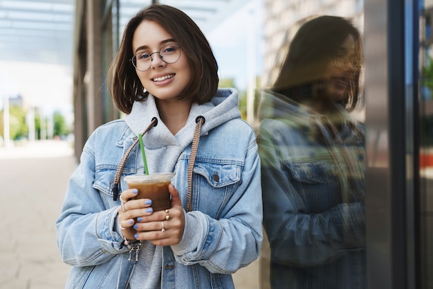 Chica joven y atractiva con gafas, chaqueta vaquera, caminar informal en la ciudad, disfrutar los fines de semana, beber café con leche, apoyarse en la pared del edificio y sonreír a cámara con expresión feliz y relajada.