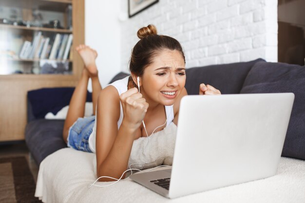 Chica joven atractiva que pone en el sofá en la sala de estar con la computadora portátil.