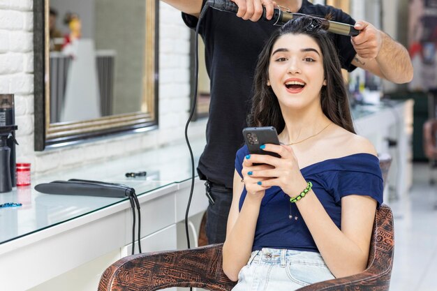 Chica joven atractiva sosteniendo el teléfono y sonriendo a la cámara en la barbería Foto de alta calidad