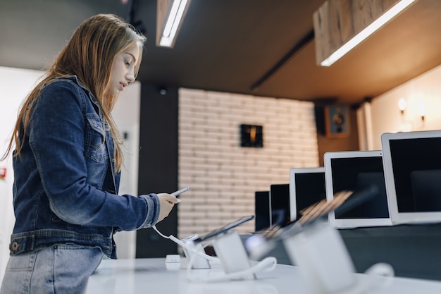 Chica joven atractiva en la tienda de electrónica se encuentra en el escritorio y prueba el teléfono