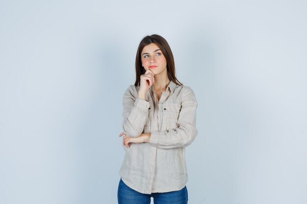 Chica joven con la barbilla inclinada en la mano, mirando a otro lado con camisa beige, jeans y mirando pensativo, vista frontal.