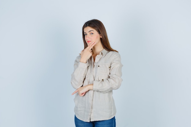 Chica joven con la barbilla inclinada en la mano, pensando en algo, apuntando hacia abajo con el dedo índice en camisa beige, jeans y mirando pensativo, vista frontal.