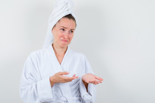 Chica joven en bata de baño blanca, toalla extendiendo las palmas mientras llora y parece impotente, vista frontal.