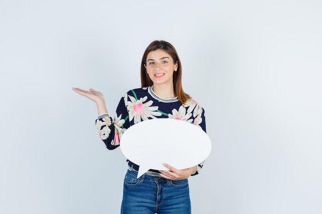 Chica joven en blusa floral, jeans extendiendo la palma y mirando feliz, vista frontal.