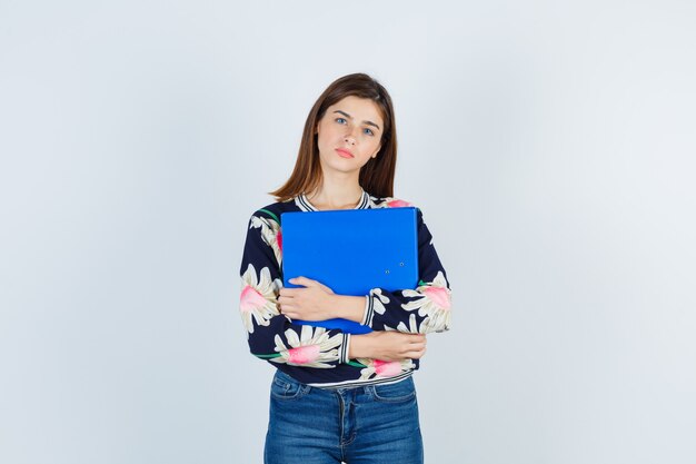 Chica joven en blusa floral, jeans sosteniendo una carpeta y mirando nostálgica, vista frontal.