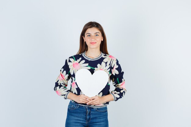 Chica joven en blusa floral, jeans sosteniendo un cartel de papel y mirando satisfecho, vista frontal.