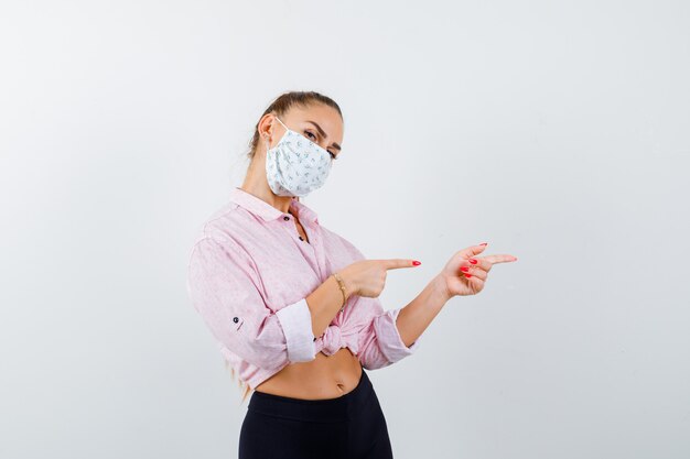 Chica joven en blusa rosa, pantalón negro, máscara apuntando hacia la derecha con los dedos índices y mirando seria, vista frontal.