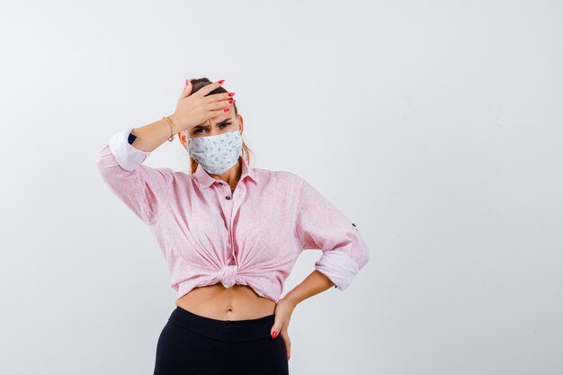 Chica joven en blusa rosa, pantalón negro, máscara poniendo la mano en la cabeza, sosteniendo una mano en la cintura y mirando enojado, vista frontal.