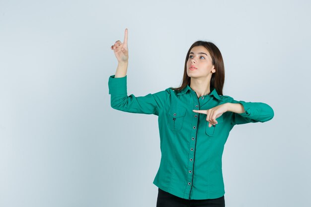 Chica joven en blusa verde, pantalón negro apuntando hacia arriba y hacia la izquierda con el dedo índice y mirando enfocado, vista frontal.