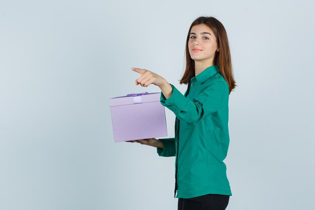 Chica joven en blusa verde, pantalón negro con caja de regalo, apuntando hacia afuera con el dedo índice y mirando alegre, vista frontal.