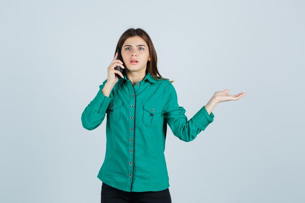 Foto gratuita chica joven en blusa verde, pantalón negro hablando por teléfono, extendiendo la palma hacia un lado y mirando sorprendido, vista frontal.