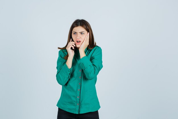 Chica joven en blusa verde, pantalón negro hablando por teléfono, sosteniendo la mano en la mejilla y mirando sorprendido, vista frontal.