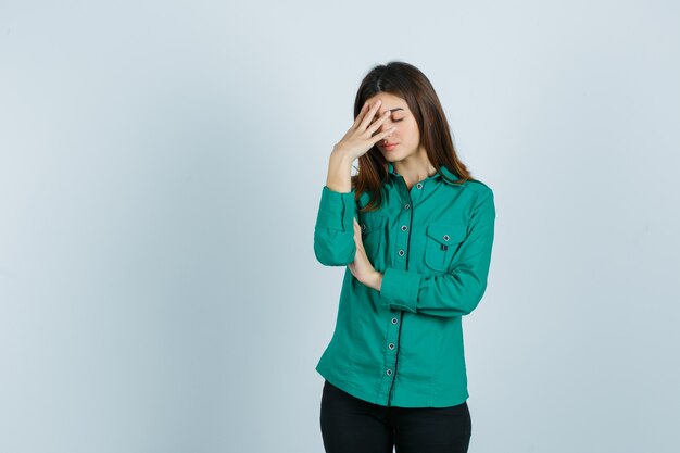 Chica joven en blusa verde, pantalón negro poniendo la mano en la frente y mirando exhausto, vista frontal.