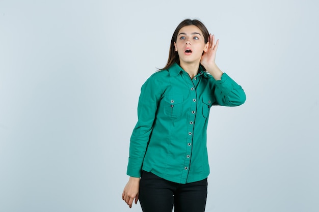 Chica joven en blusa verde, pantalón negro sosteniendo la mano cerca de la oreja para escuchar algo y mirando enfocado, vista frontal.