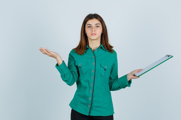 Chica joven en blusa verde, pantalón negro sosteniendo el portapapeles, extendiendo la palma hacia un lado y mirando seria, vista frontal.