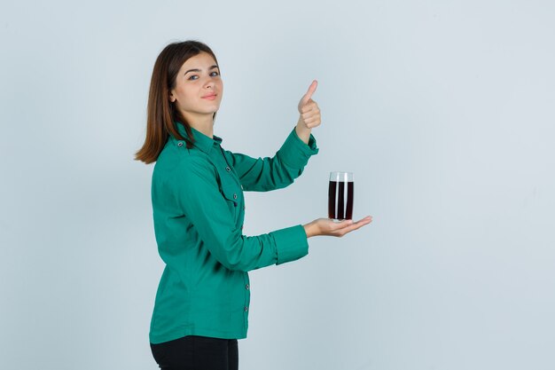 Chica joven en blusa verde, pantalón negro sosteniendo un vaso de líquido negro, mostrando el pulgar hacia arriba y mirando alegre, vista frontal.