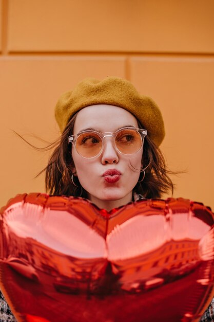 Chica joven en boina amarilla envía beso Mujer escondida detrás de globo rojo en forma de corazón